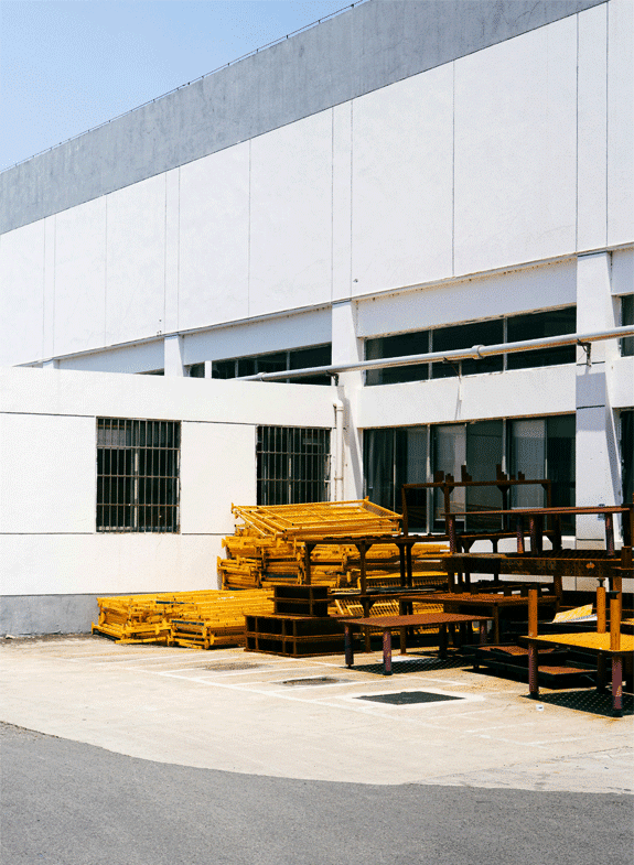 Exterior view of a large, white industrial or warehouse building on a sunny day, with stacks of yellow and rusty brown metal frames, racks, or scaffolding stored in the parking lot in the foreground.