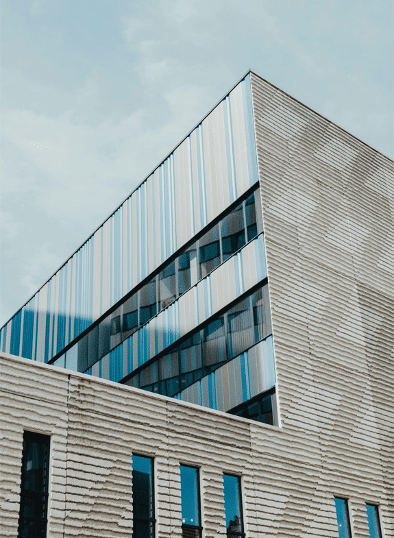 Architectural close-up of a modern building facade featuring a sharp corner. One section has textured, light-colored horizontal siding, and the upper section has vertical striped siding in shades of light blue, white, and silver, reflecting the sky.